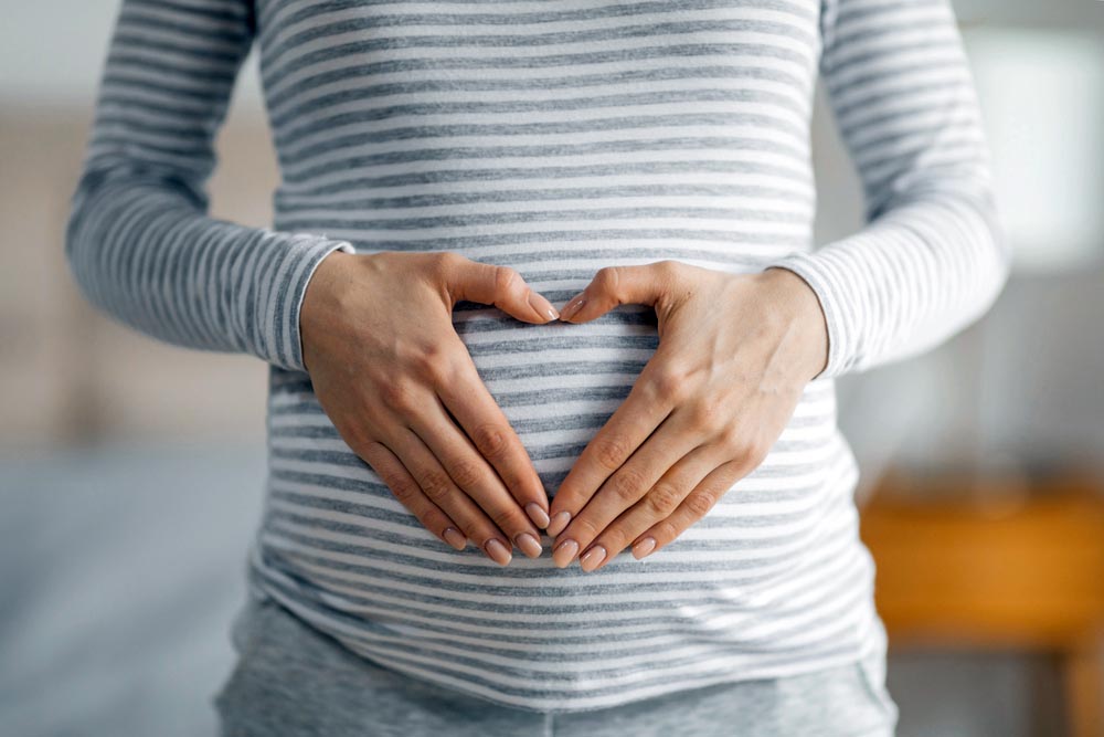 Close-up of a pregnant woman forming a heart shape with her hands on her striped shirt covering the belly