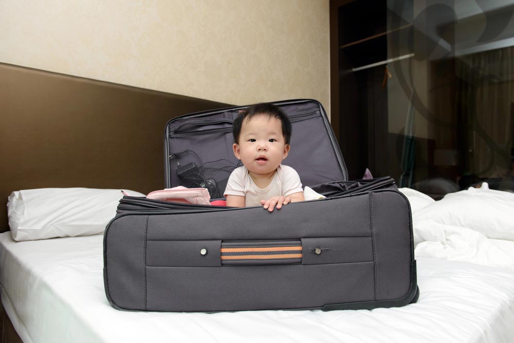 Asian baby sitting in travel suitcase placed on hotel double bed